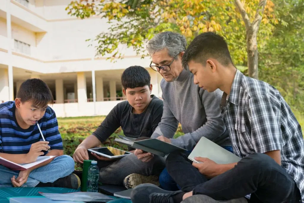 An educator and three students sitting on the grass outside a school, reading and talking together